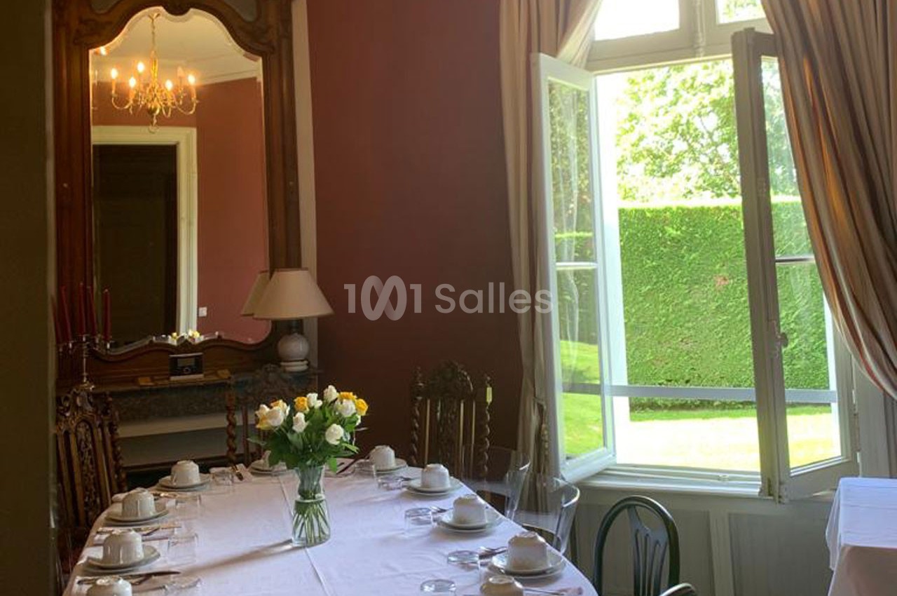 Salle à manger lumineuse avec table dressée, miroir ancien, murs rouges et grande fenêtre ouverte sur un jardin.