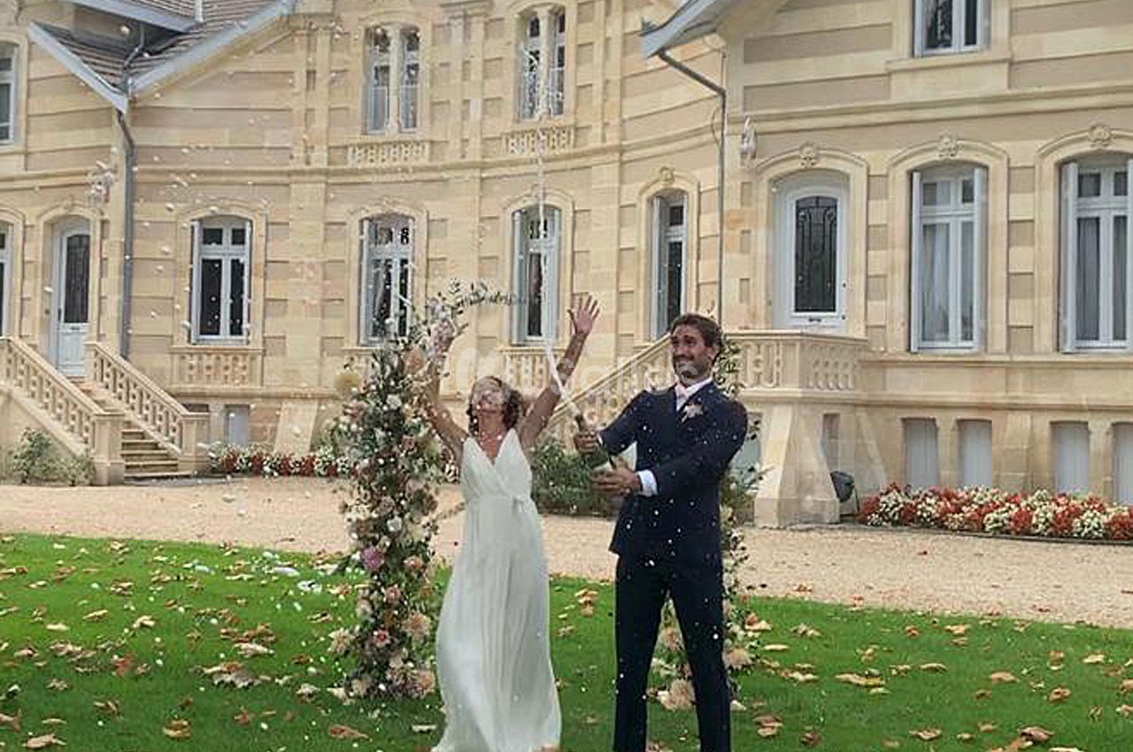 Un couple célèbre un mariage devant une grande bâtisse, entouré de fleurs et de pétales tombant.