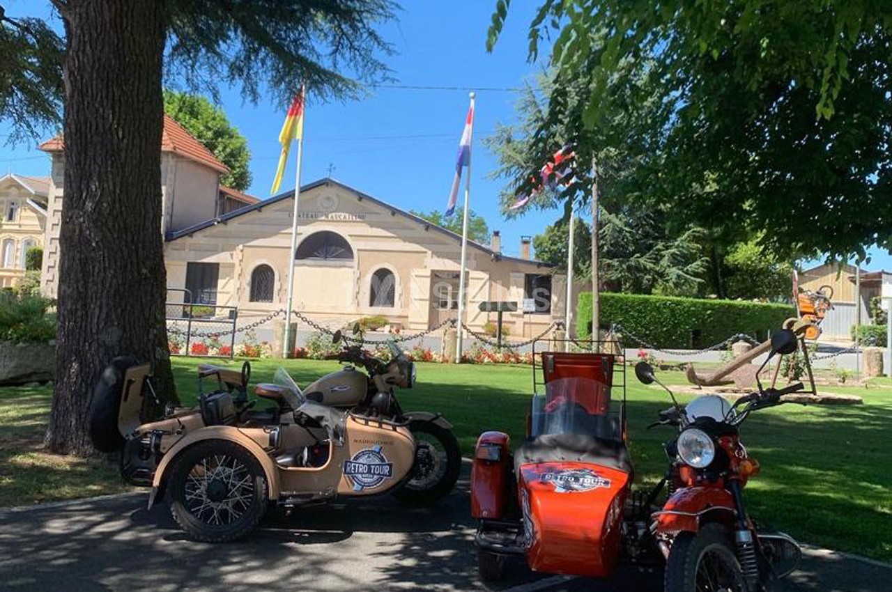 Deux motos avec side-cars stationnées dans un parc ombragé devant un bâtiment ancien et des drapeaux.