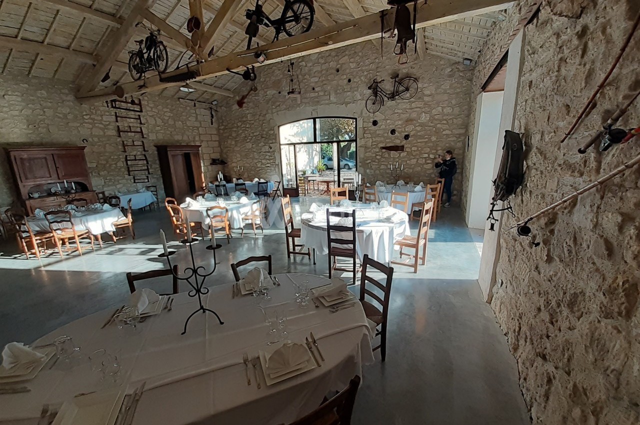 Salle en pierre avec tables dressées pour un repas, éclairée par la lumière naturelle entrant par une grande baie vitrée.