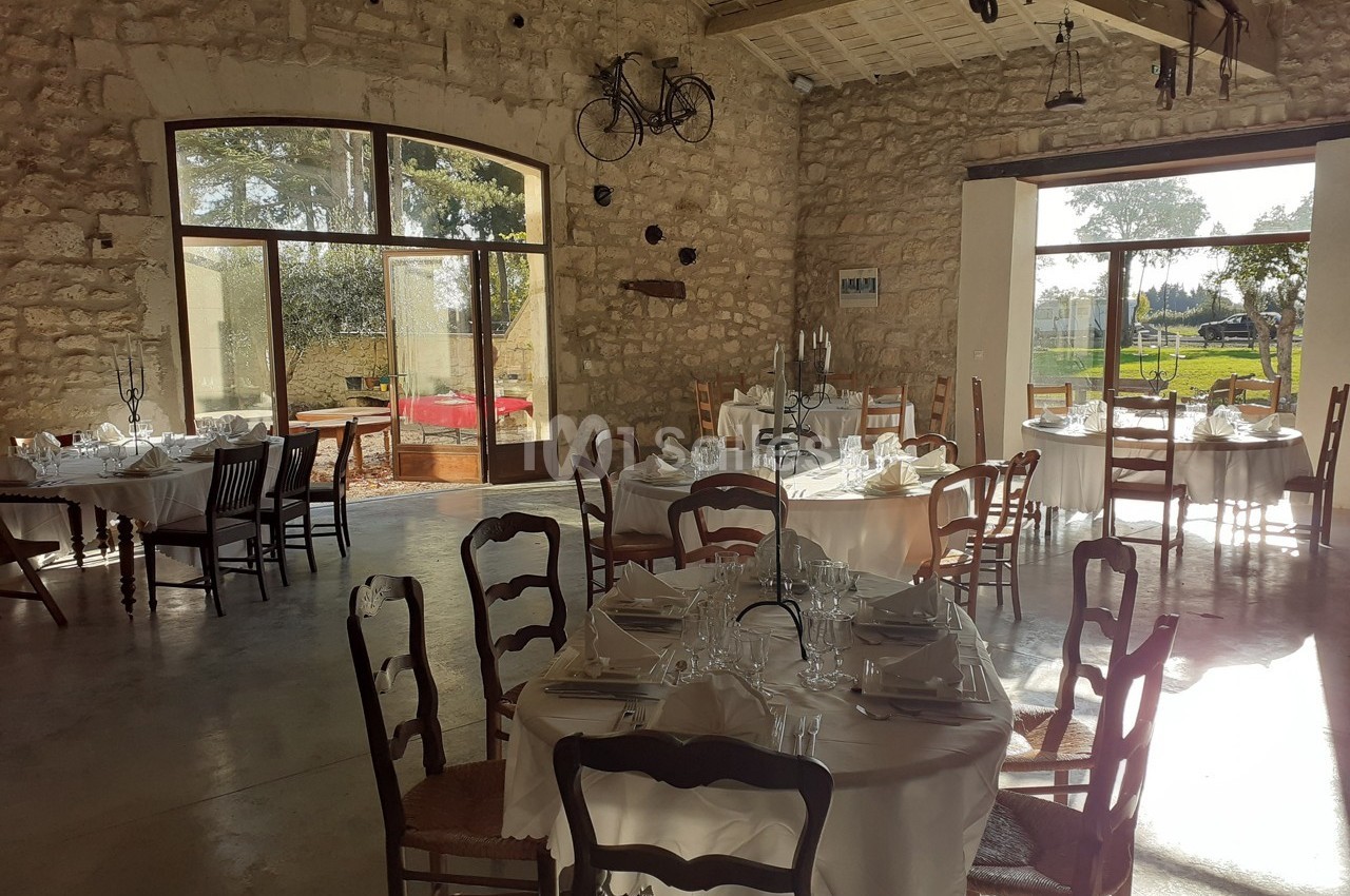 Salle de réception lumineuse avec des tables rondes dressées, des chaises en bois et des murs en pierre apparente.