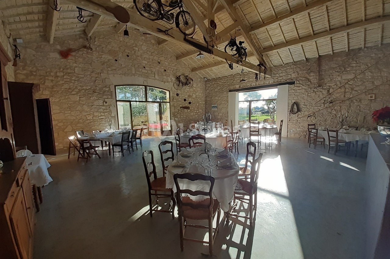 Salle lumineuse avec murs en pierre, tables dressées et décorations suspendues, donnant sur un extérieur ensoleillé.