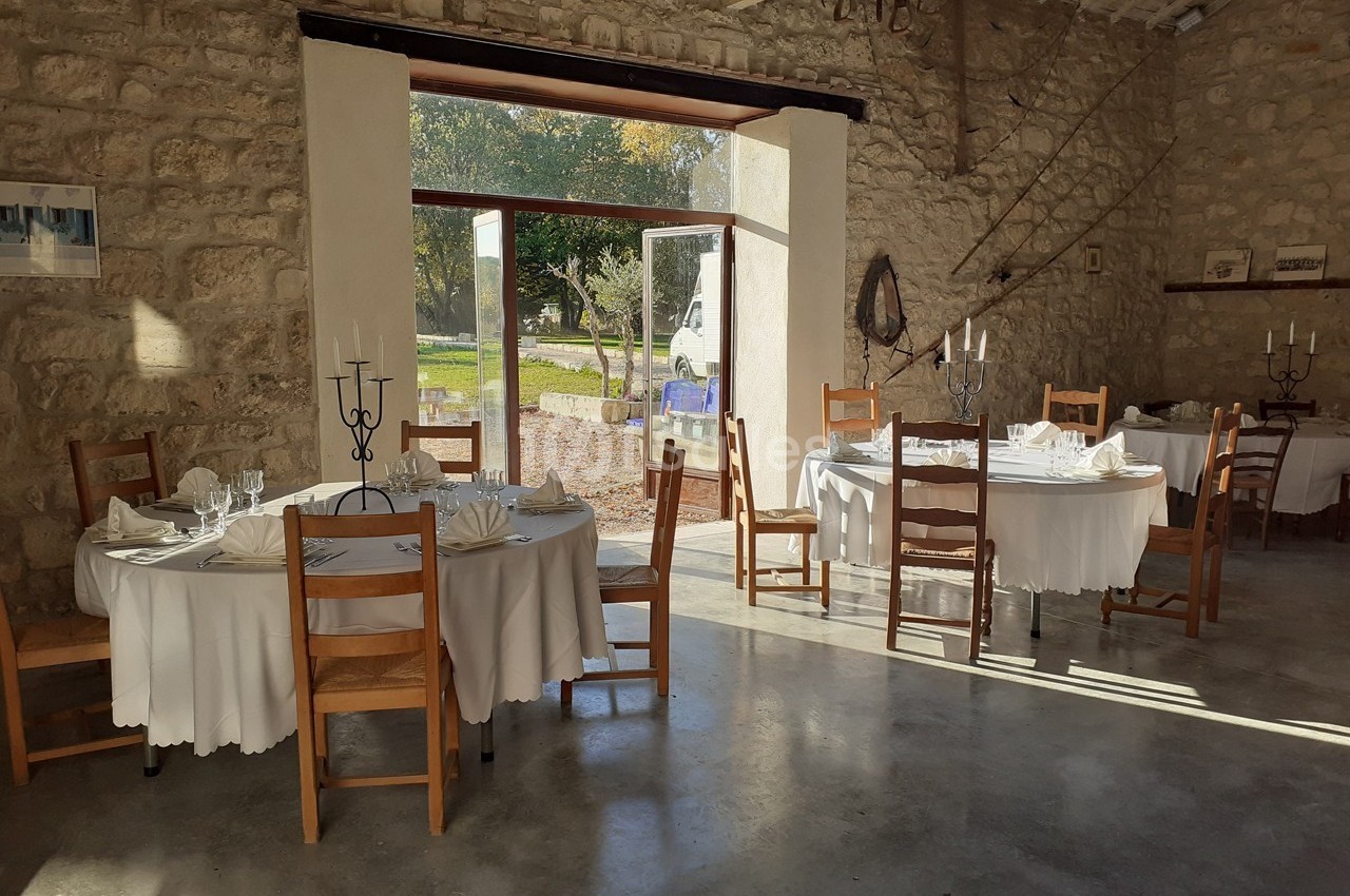 Salle en pierre lumineuse avec tables rondes dressées, nappes blanches et chaises en bois, donnant sur un extérieur…