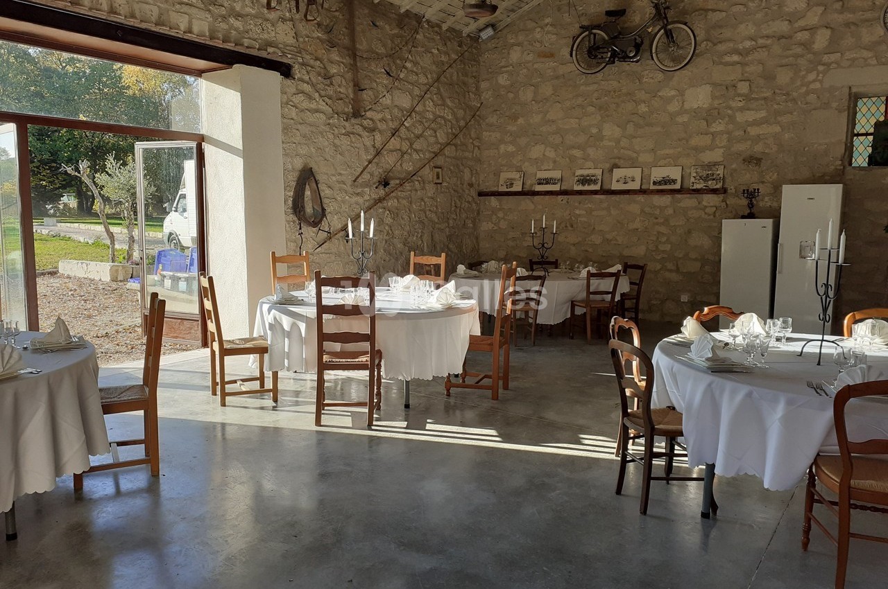 Salle en pierre avec grandes baies vitrées, tables rondes dressées avec nappes blanches et chandeliers.