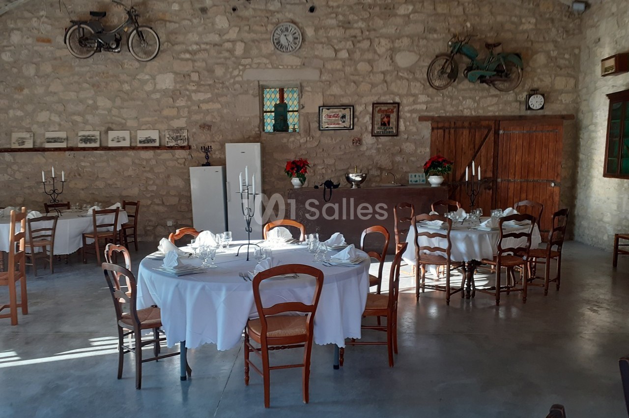 Salle en pierre avec tables rondes dressées, chaises en bois, décorations murales vintage et lumière naturelle.