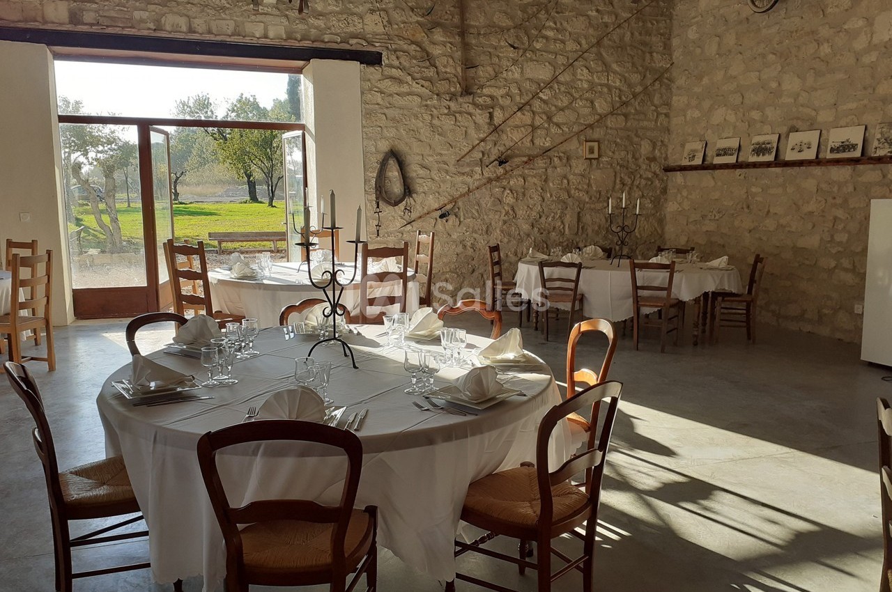 Salle en pierre lumineuse avec tables rondes dressées, nappes blanches, chaises en bois et vue sur un jardin verdoyant.