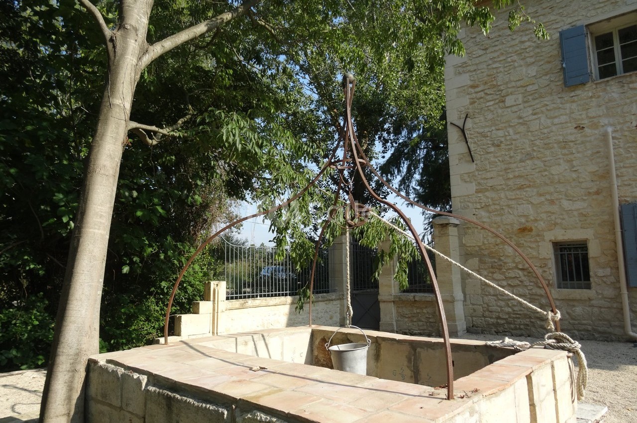 Puits en pierre avec une structure métallique et un seau suspendu, situé près d'une maison en pierre et d'un arbre.