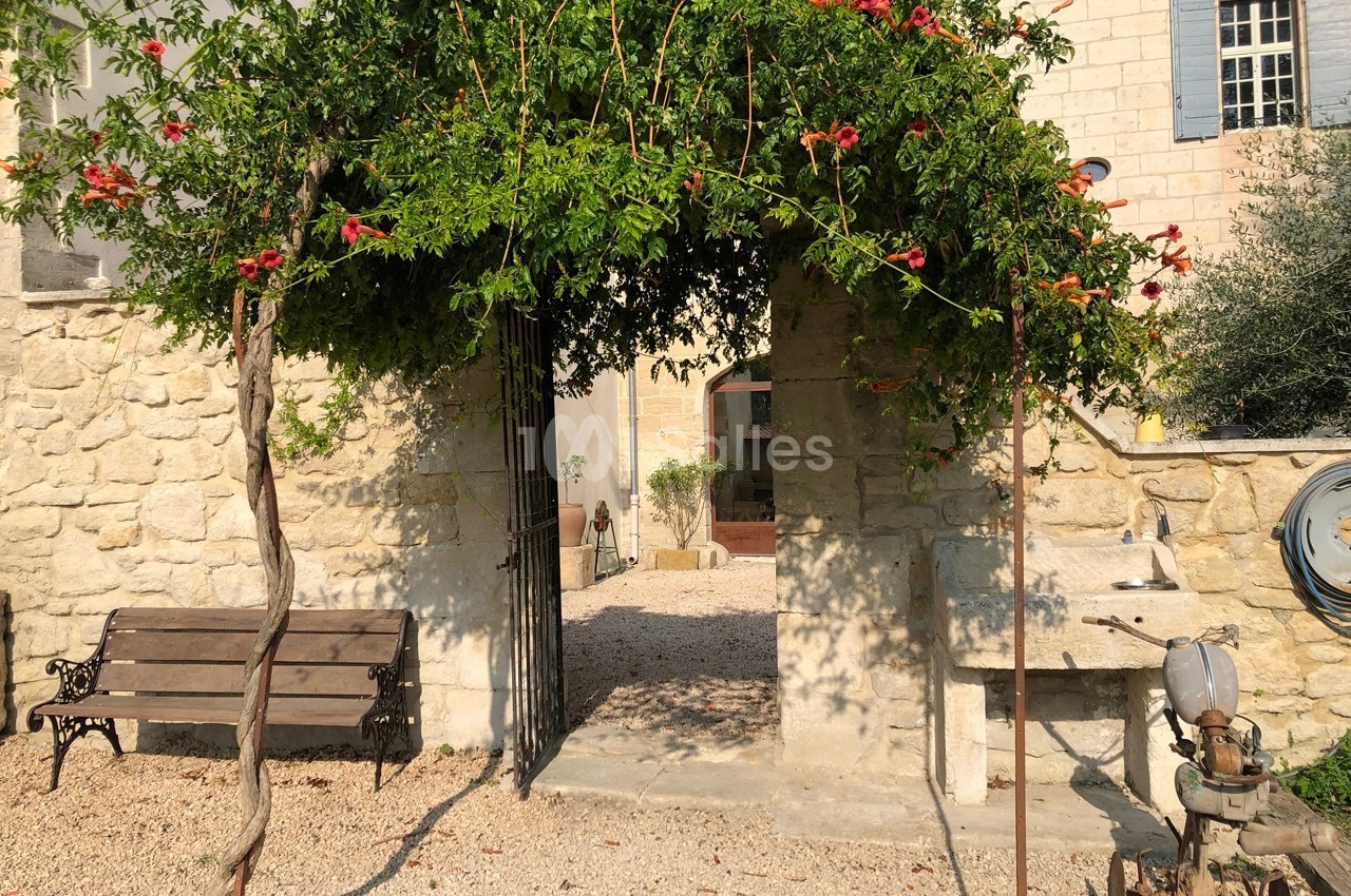 Arche en pierre recouverte de végétation fleurie, donnant sur une cour en gravier avec un banc et des éléments en pierre.
