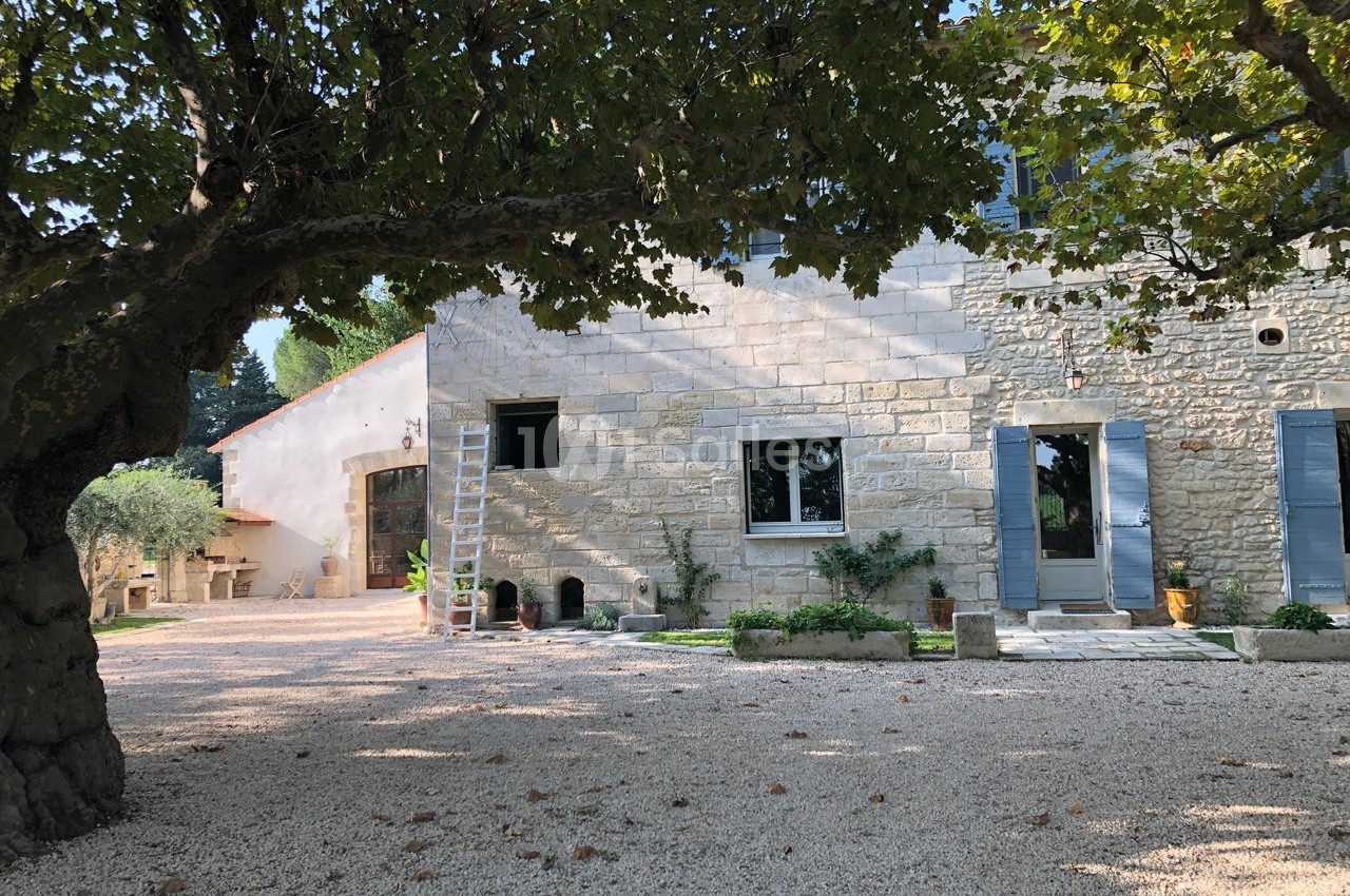 Façade en pierre d'une maison avec volets bleus, entourée d'arbres et d'une cour en gravier.