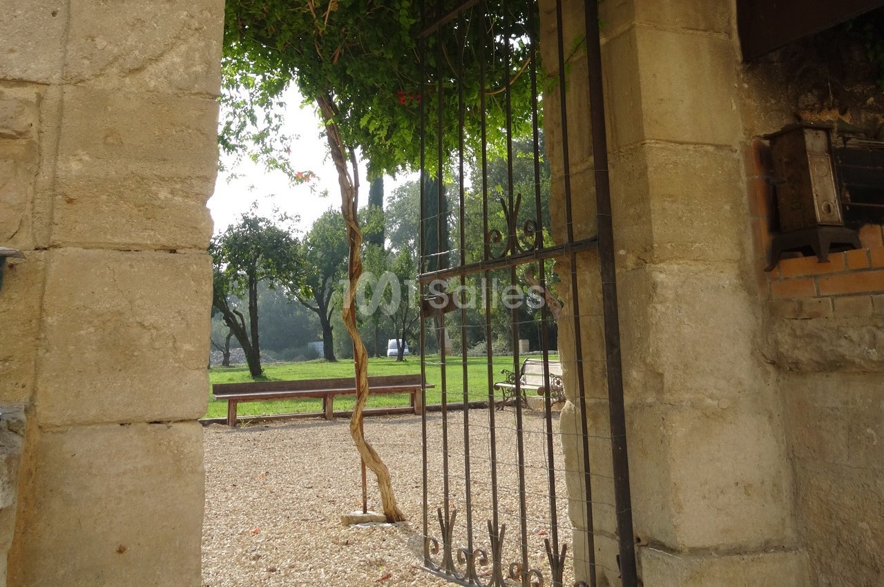 Portail en fer forgé ouvert sur un jardin avec bancs, arbres et lumière naturelle filtrée par les feuillages.