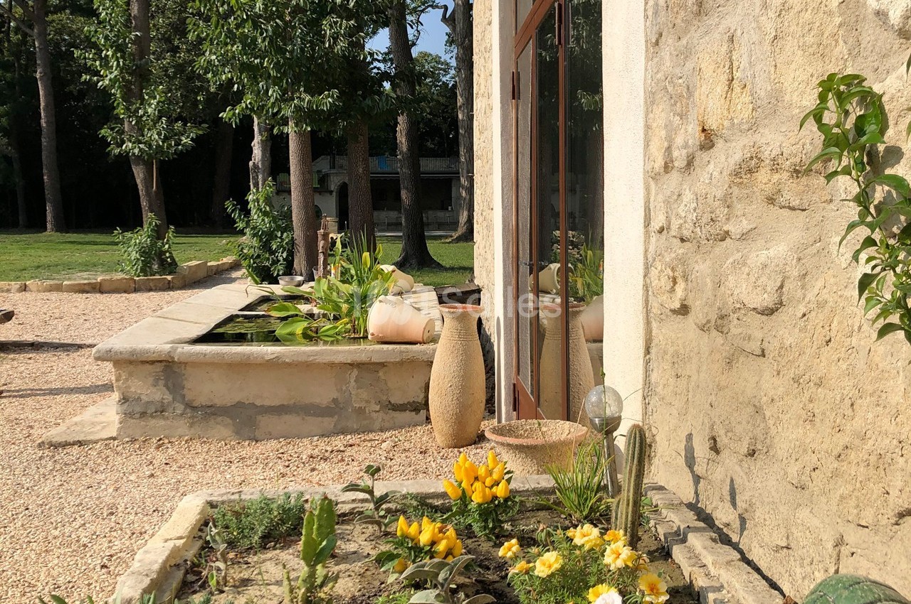 Jardin avec parterre fleuri, poteries décoratives et bassin près d'une façade en pierre sous un ciel ensoleillé.