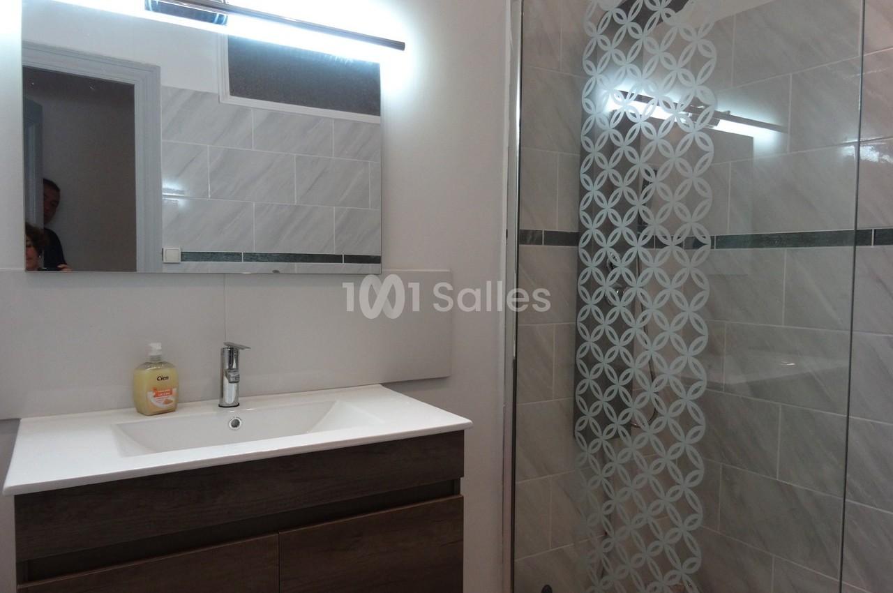 Salle de bain moderne avec lavabo, meuble en bois sombre, miroir éclairé et douche avec paroi en verre décorée.