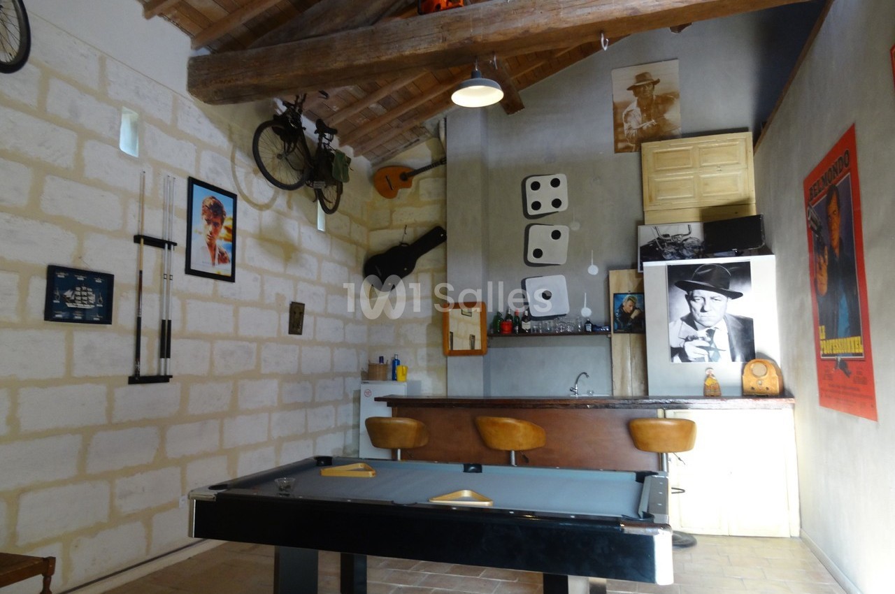 Salle de loisirs avec billard, bar, décorations murales variées et plafond en poutres apparentes.