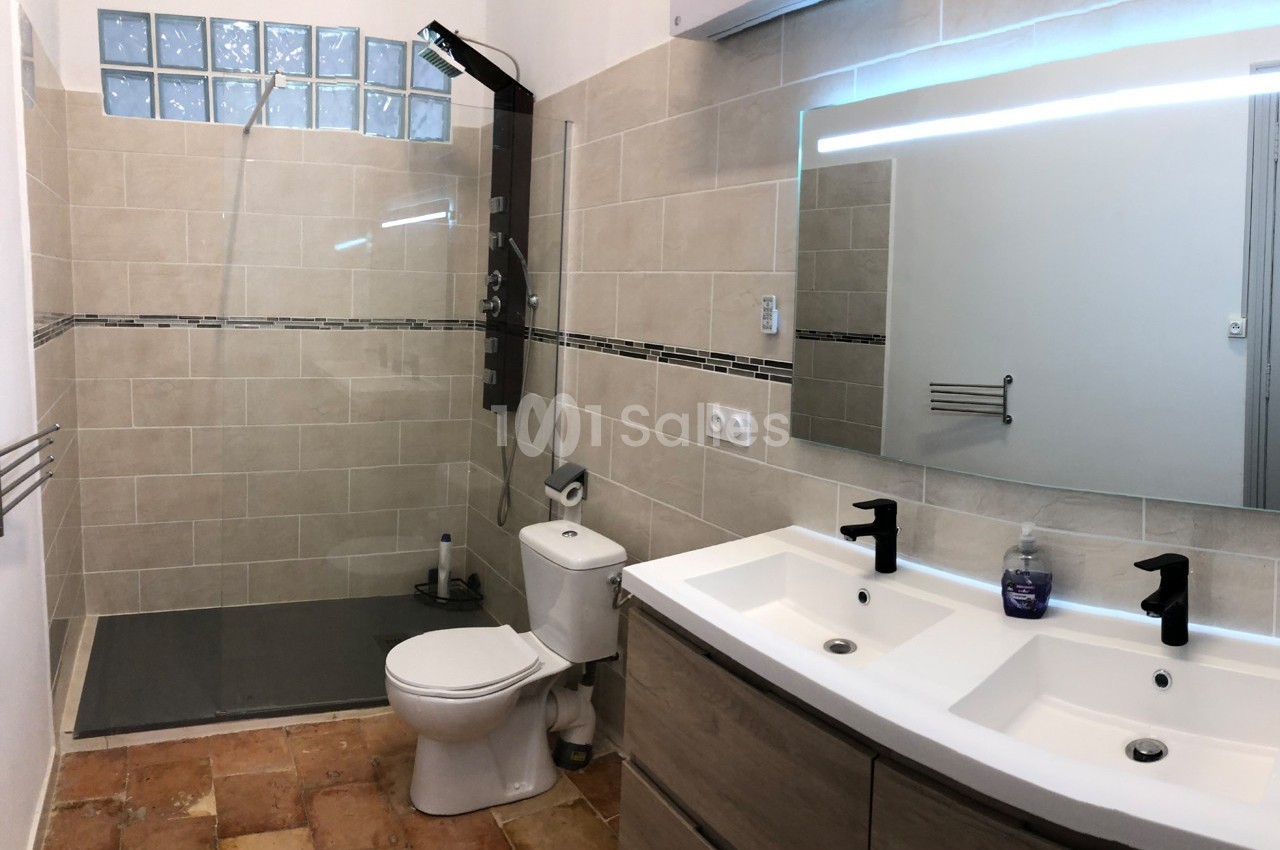 Salle de bain avec douche à l'italienne, lavabo double vasque, miroir éclairé et toilettes sur carrelage en terre cuite.
