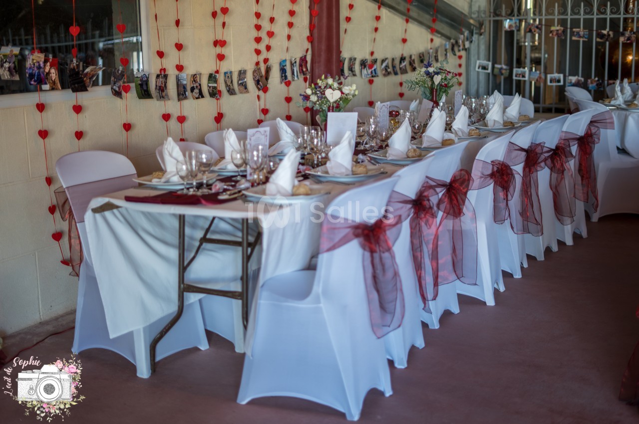 Table décorée pour un événement, avec nappes blanches, serviettes pliées, rubans rouges et guirlandes de cœurs en arrière…