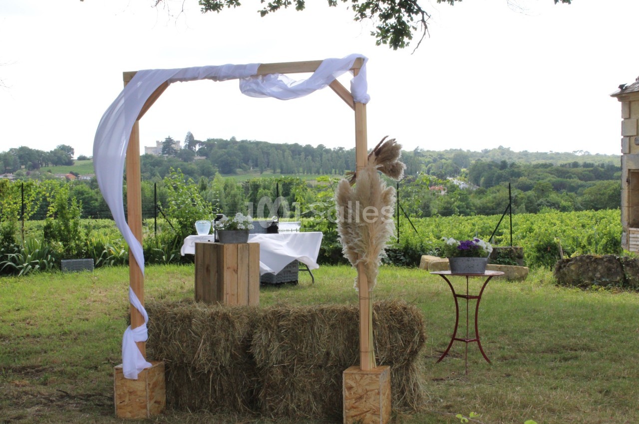 Arche en bois décorée de voiles blancs et de pampas, installée dans un champ avec une table dressée en arrière-plan.