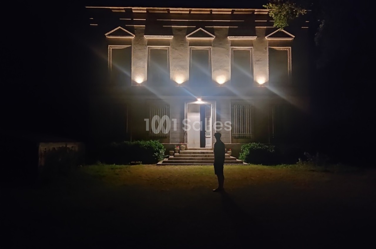 Silhouette d'une personne debout devant une maison éclairée la nuit, entourée de végétation.