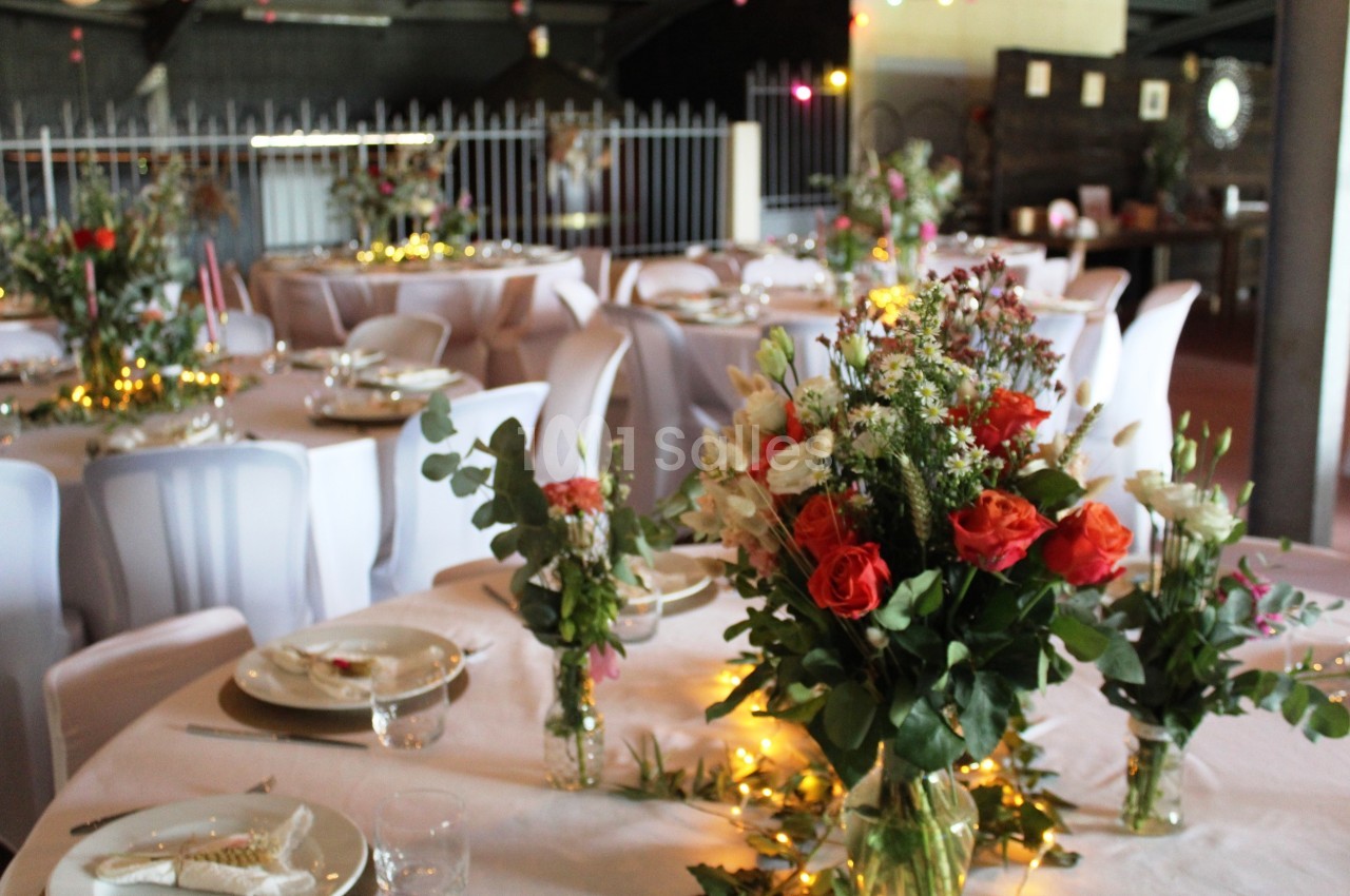 Salle décorée pour un événement avec des tables rondes, nappes blanches, bouquets de fleurs et guirlandes lumineuses.