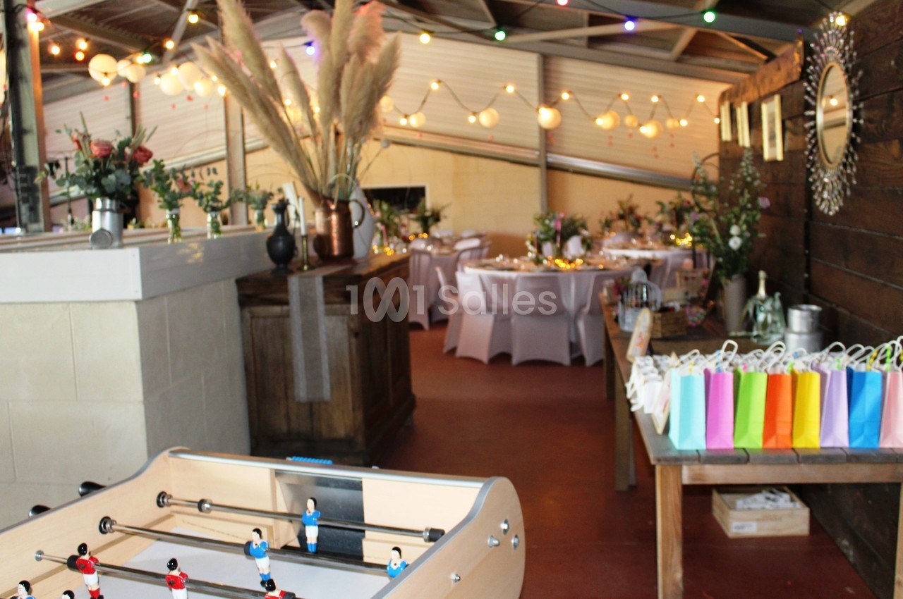 Salle décorée pour un événement avec des tables dressées, des lumières suspendues et un baby-foot au premier plan.