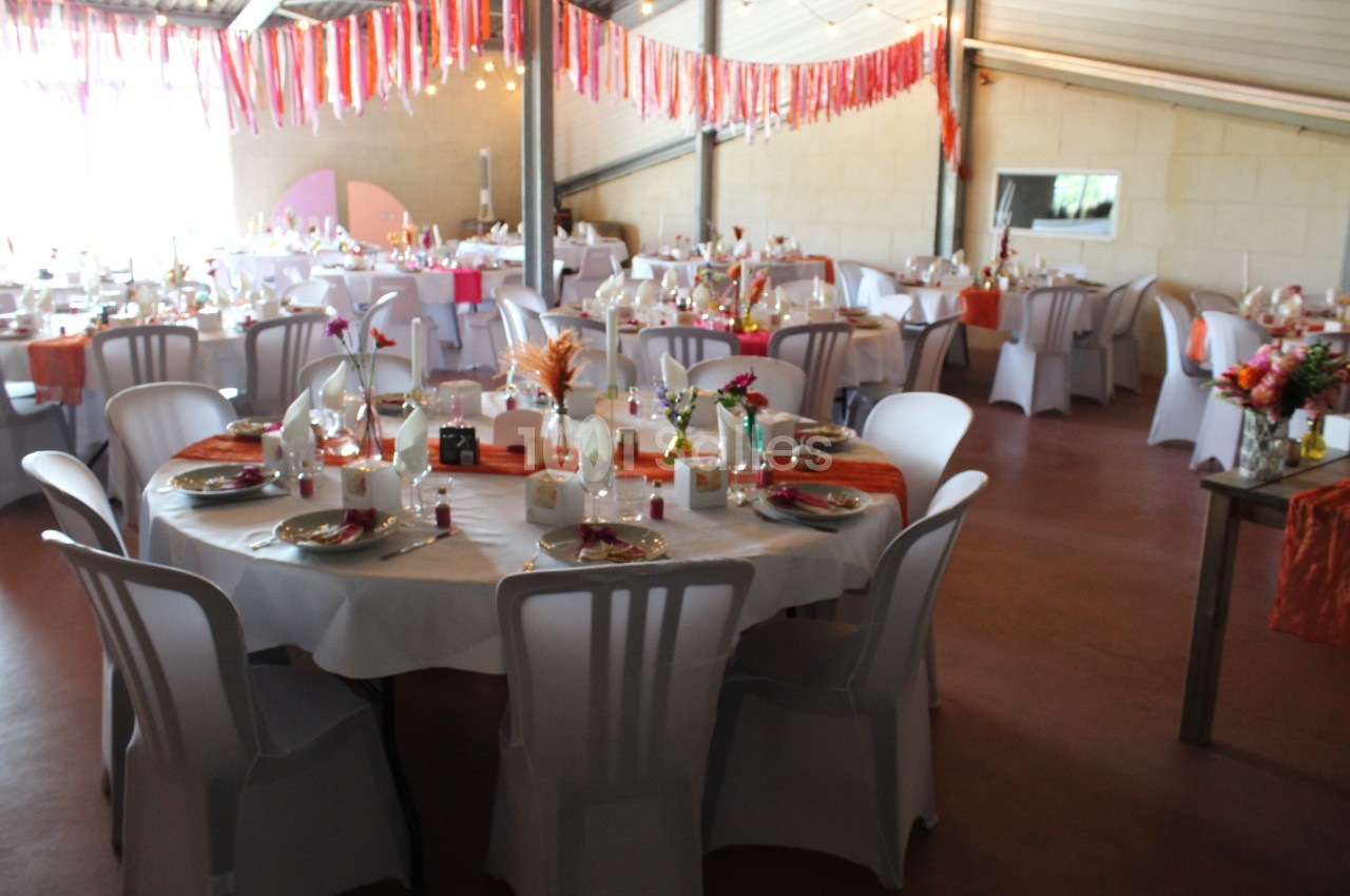Salle décorée pour un événement avec des tables rondes dressées, nappes blanches et décorations colorées suspendues.