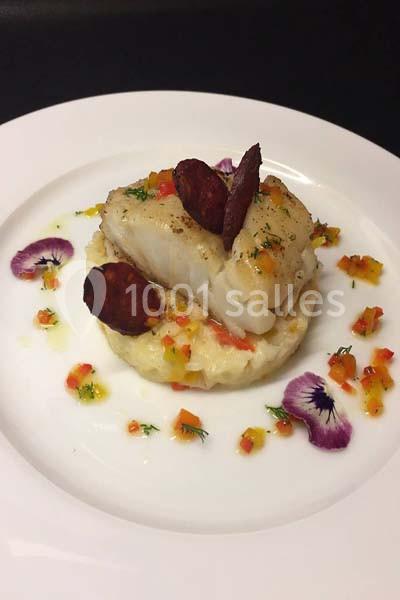Plat de poisson blanc sur purée, décoré de rondelles de chorizo, légumes colorés et fleurs comestibles.