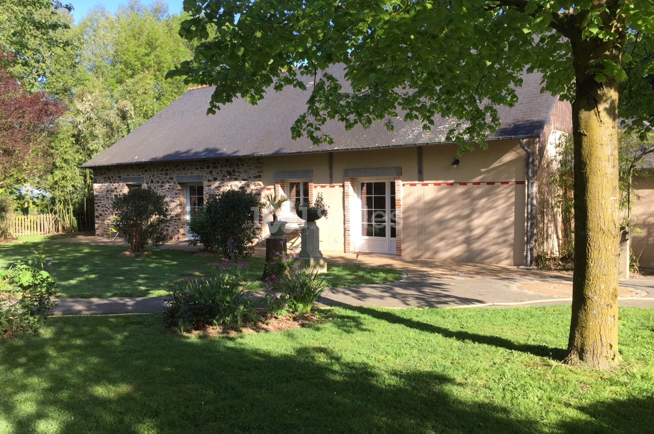 Maison en pierre avec toit en ardoise, entourée d'un jardin verdoyant et d'arbres sous un ciel dégagé.