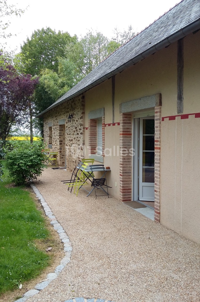 Façade d'une maison en pierre et crépi avec terrasse, mobilier de jardin et allée gravillonnée bordée de verdure.