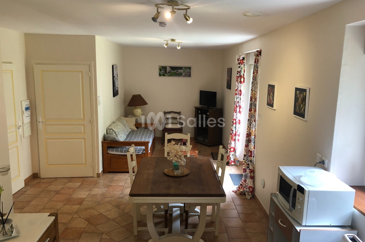 Salle de séjour lumineuse avec table à manger, canapé, télévision, et cuisine équipée visible au premier plan.