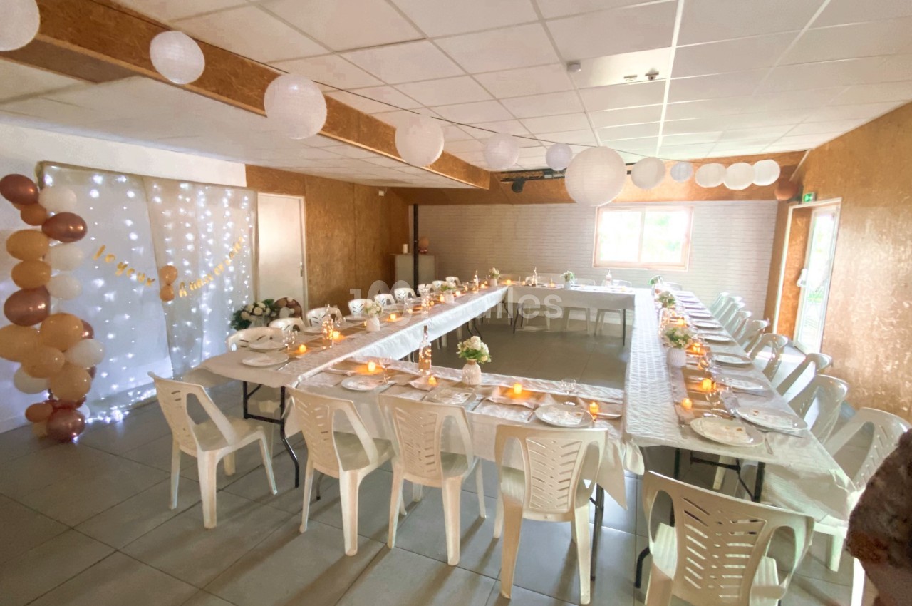 Salle décorée pour un événement, avec tables en U dressées, chaises blanches et guirlandes lumineuses en arrière-plan.