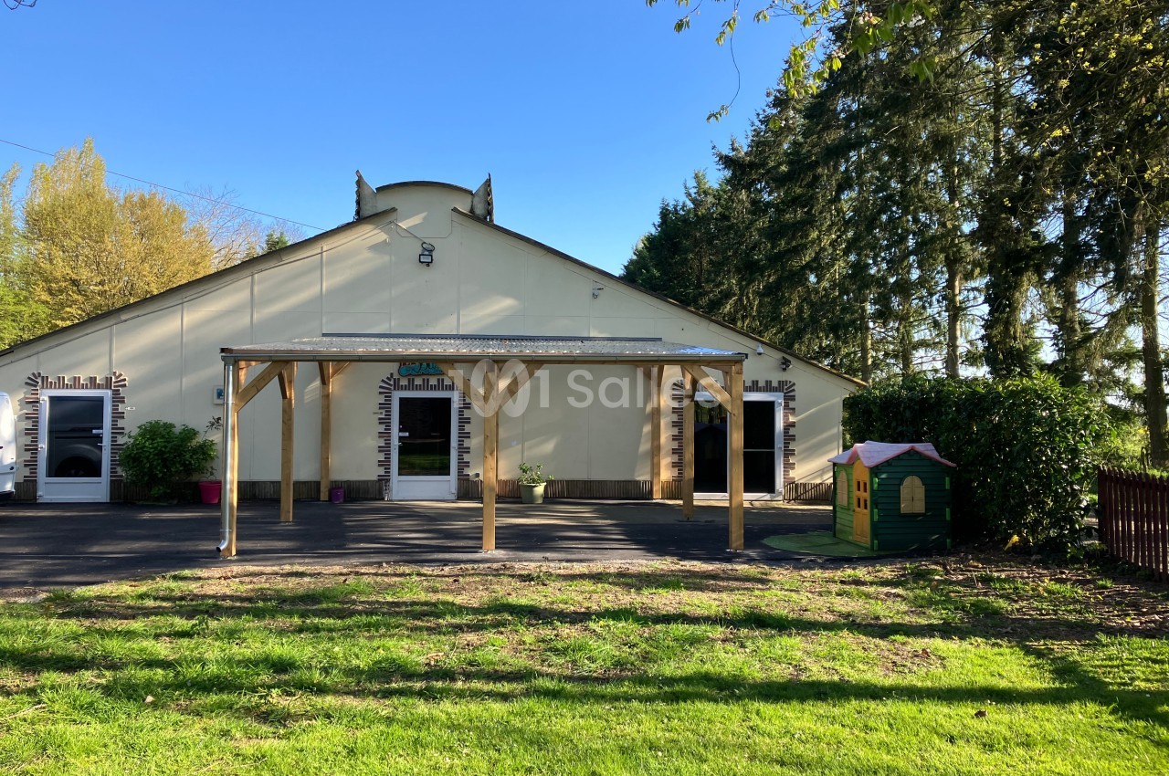 Façade d'un bâtiment clair avec une pergola en bois, entouré d'arbres et d'une pelouse avec une cabane pour enfants.