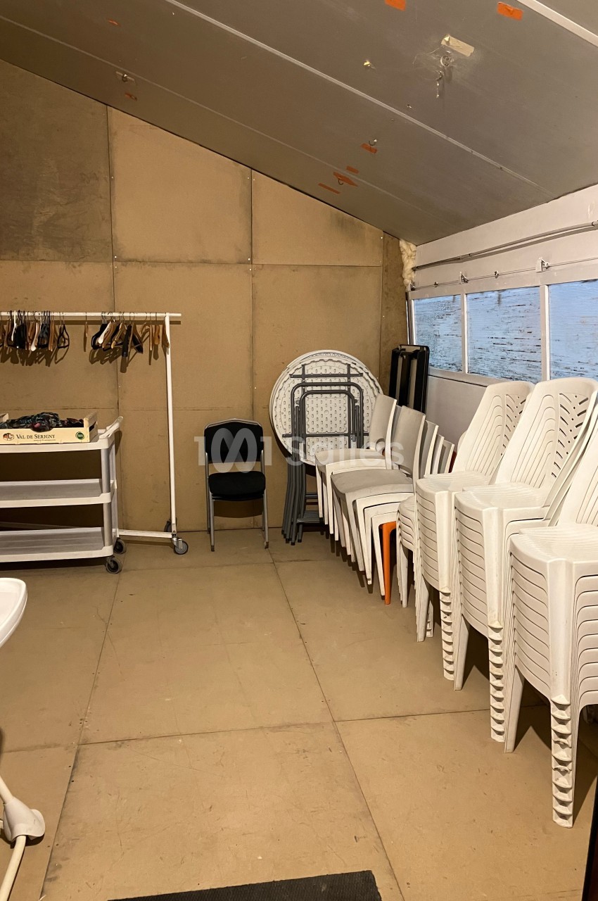 Salle de rangement avec des chaises empilées, une table pliante, un portant à cintres et un chariot à roulettes.