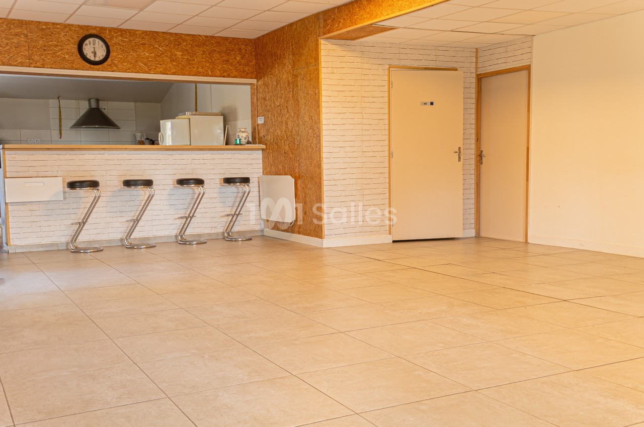 Salle lumineuse avec carrelage beige, comptoir équipé de tabourets et cuisine ouverte en arrière-plan.