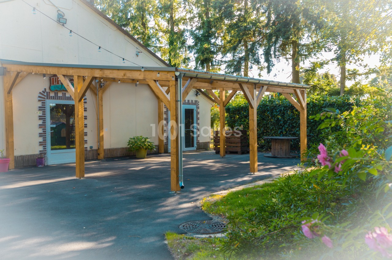 Terrasse couverte en bois devant un bâtiment beige, entourée de végétation et éclairée par une lumière naturelle.