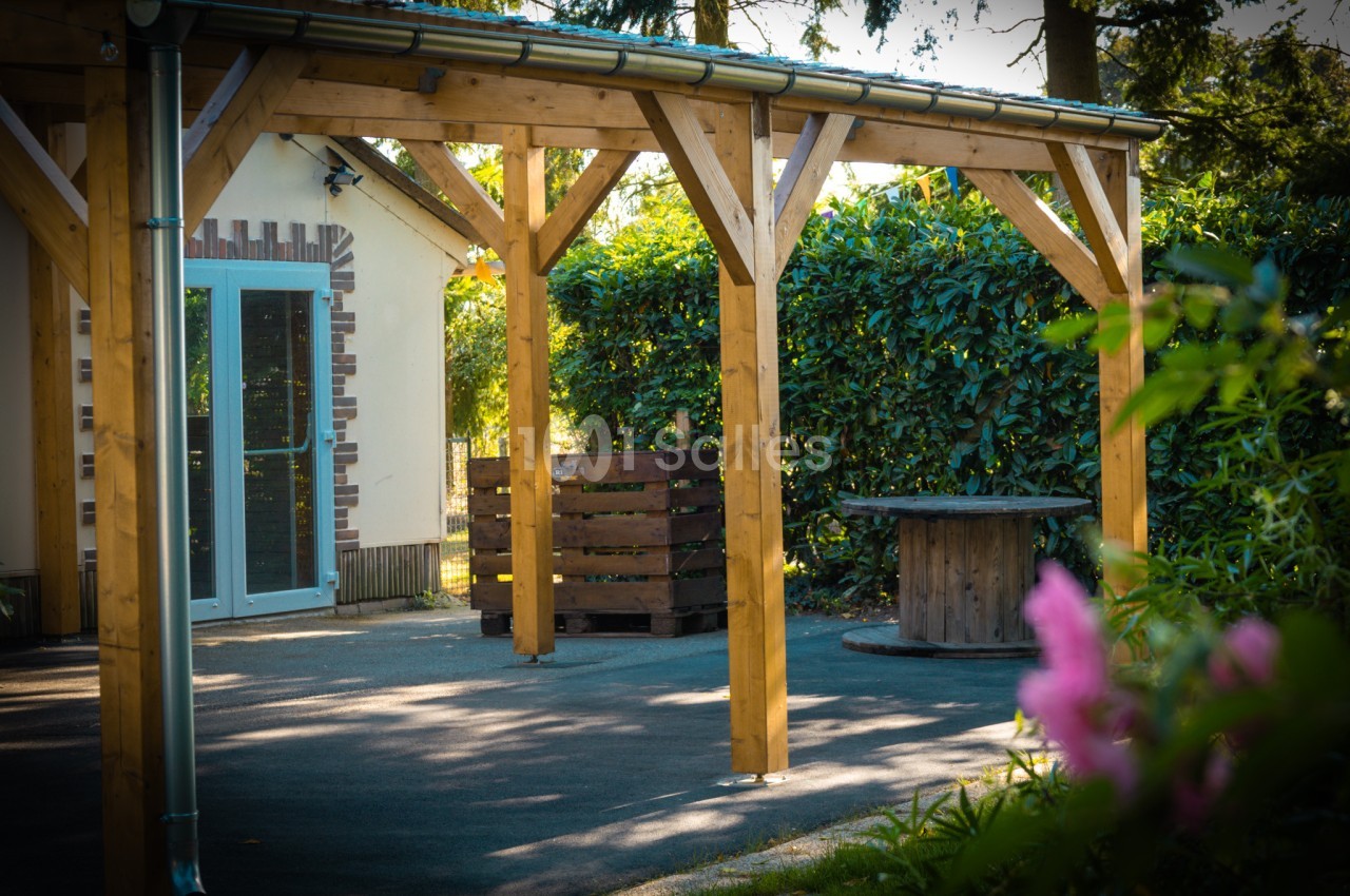 Structure en bois avec une table ronde et des palettes, située devant un bâtiment entouré de végétation.