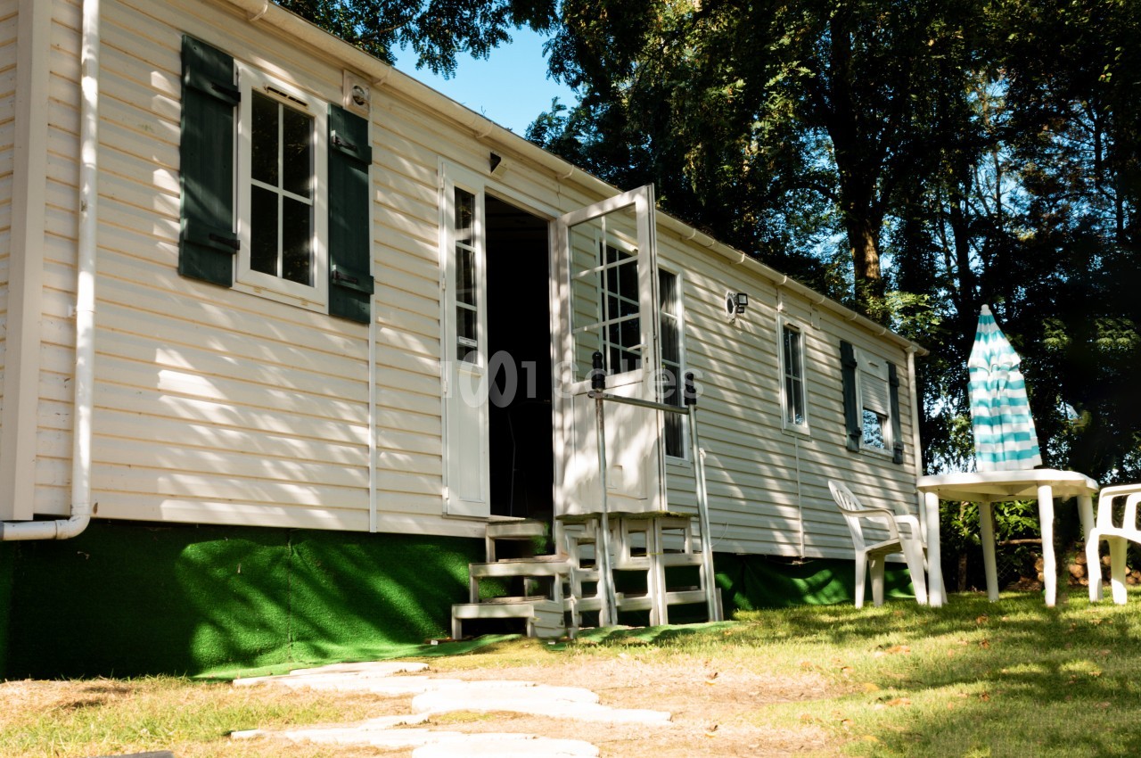 Mobil-home blanc avec porte ouverte, table et chaises en plastique sur une pelouse, entouré d'arbres.