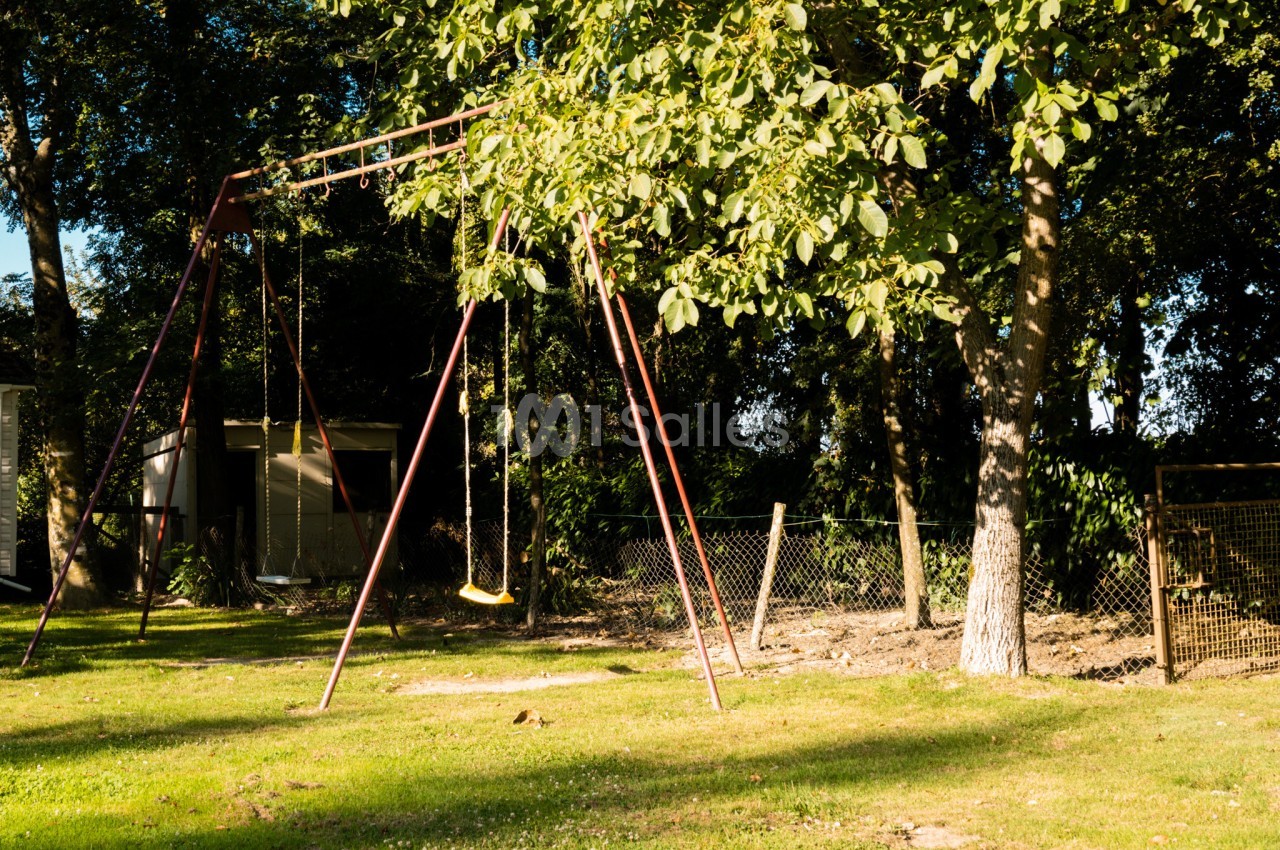 Portique avec balançoires dans un jardin ensoleillé, entouré d'arbres et de clôtures.
