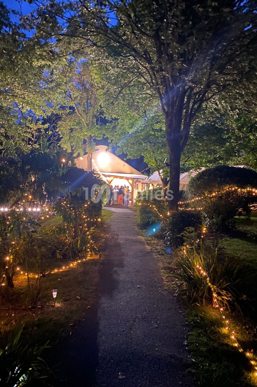 Allée éclairée par des guirlandes lumineuses menant à une maison entourée de végétation, en soirée.