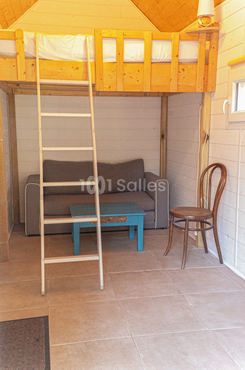 Petit espace aménagé avec un canapé gris, une table basse bleue, une chaise en bois et un lit en mezzanine accessible par…
