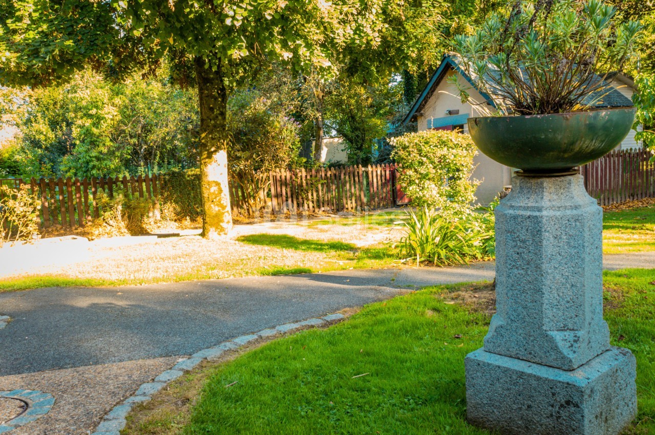 Chemin sinueux bordé de verdure et d'arbres, avec une maison en arrière-plan et un pot de fleurs sur un socle au premier…
