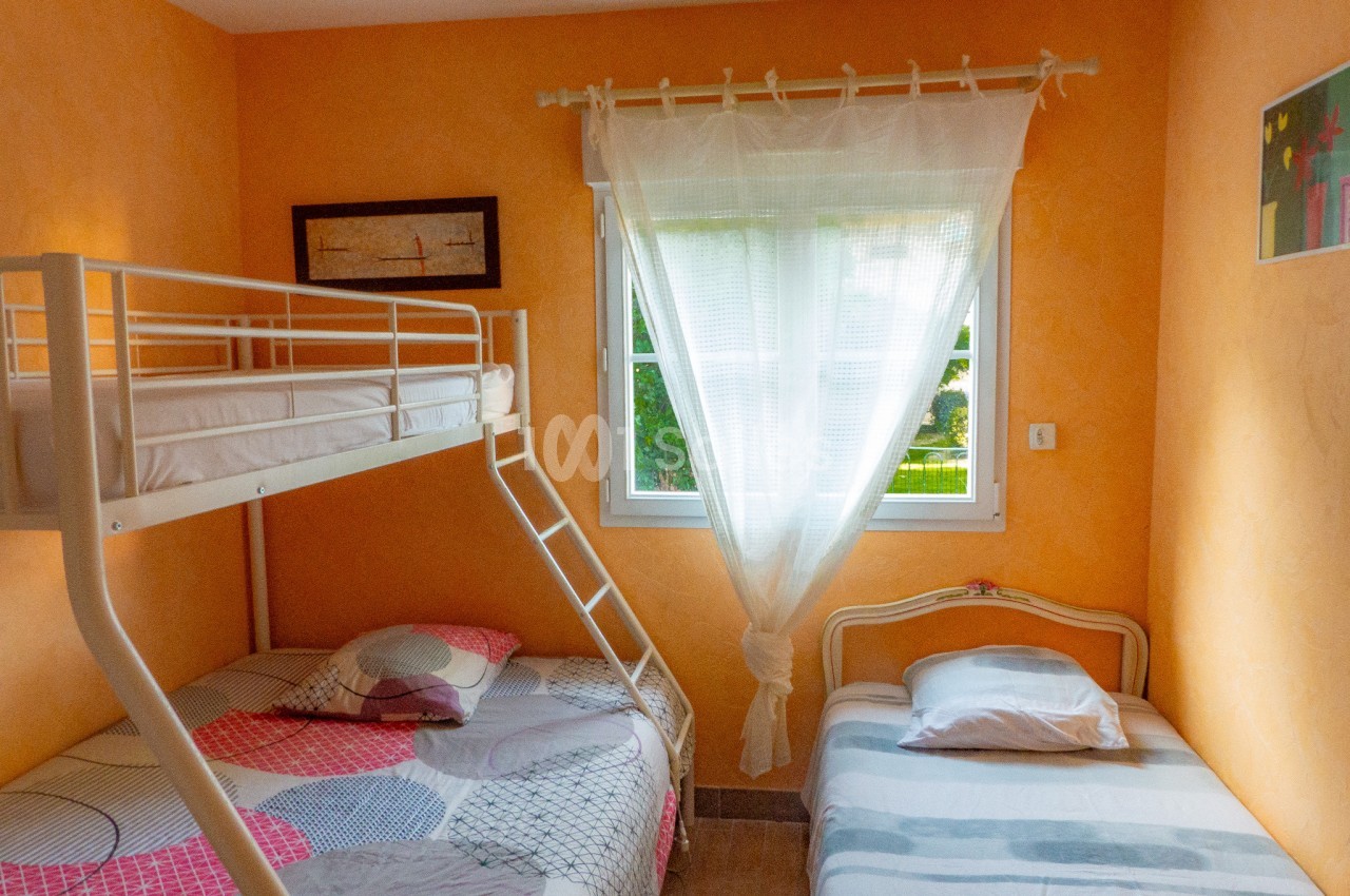 Chambre lumineuse avec murs orange, lit superposé, lit simple, fenêtre avec rideaux blancs et tableau décoratif.