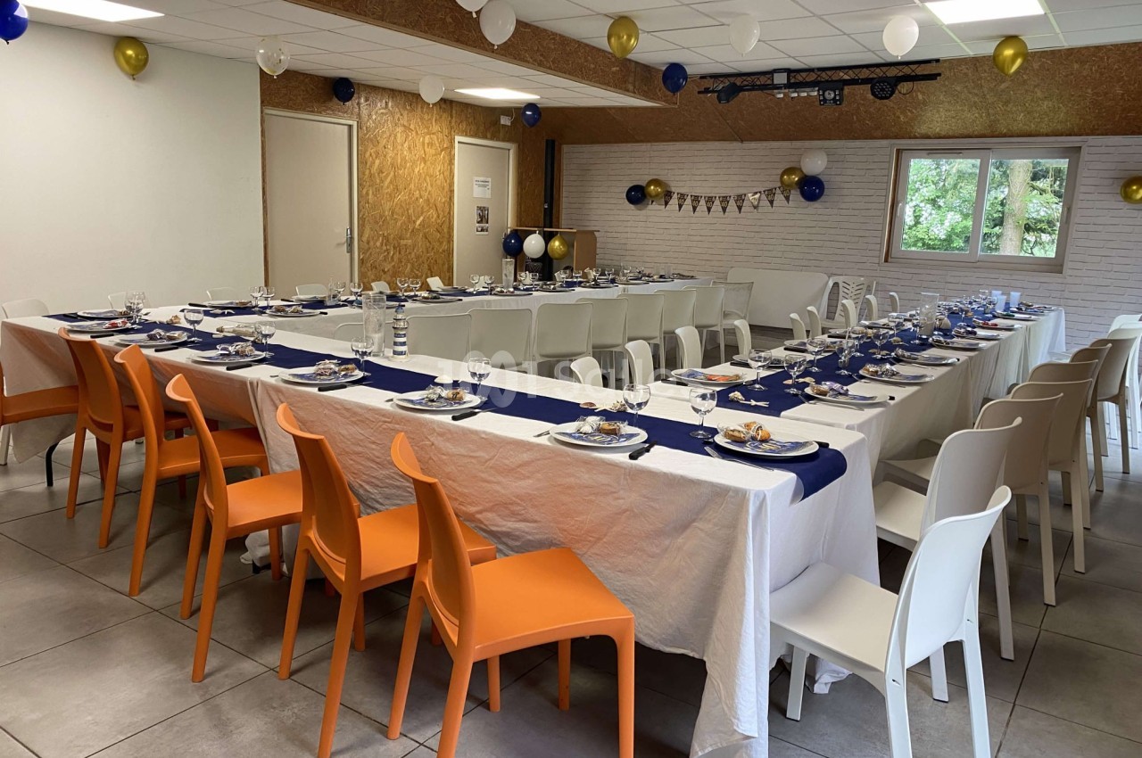Salle décorée pour un événement avec tables dressées, nappes blanches, chaises colorées et ballons suspendus.