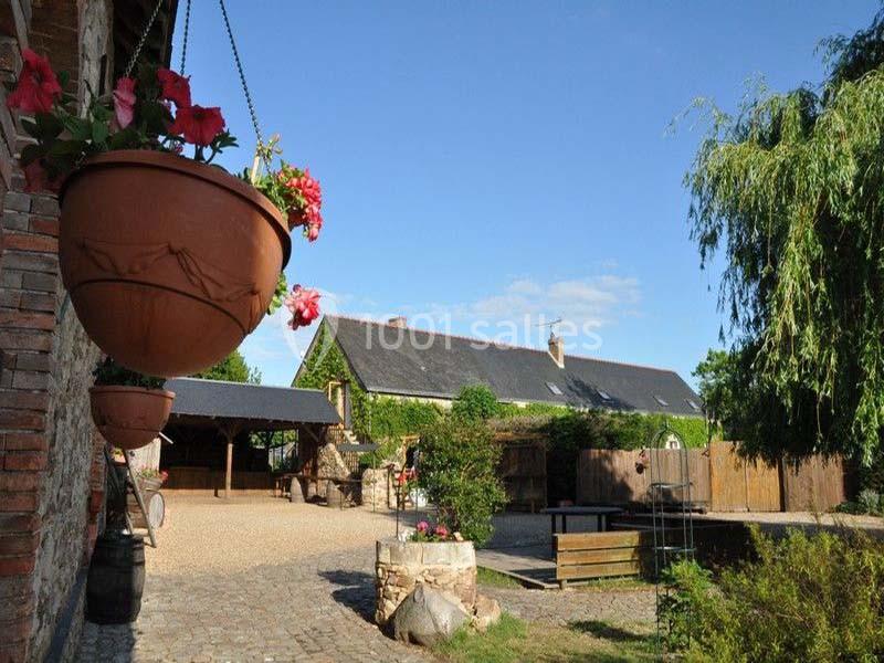 Cour pavée avec pots suspendus, puits en pierre, bâtiment en pierre et toit d'ardoise, entouré de verdure.
