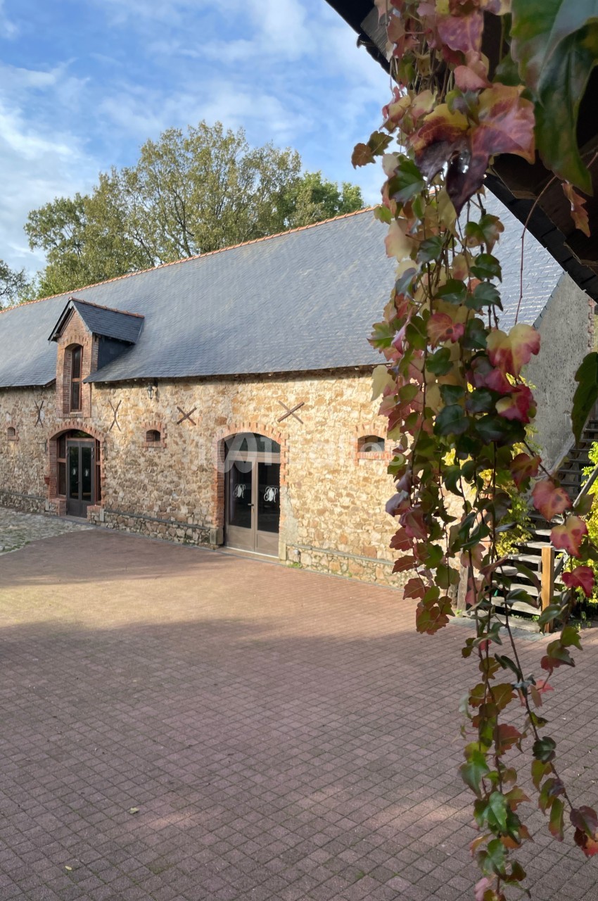 Bâtiment en pierre avec toit en ardoise, entouré de végétation, vu depuis une cour pavée.