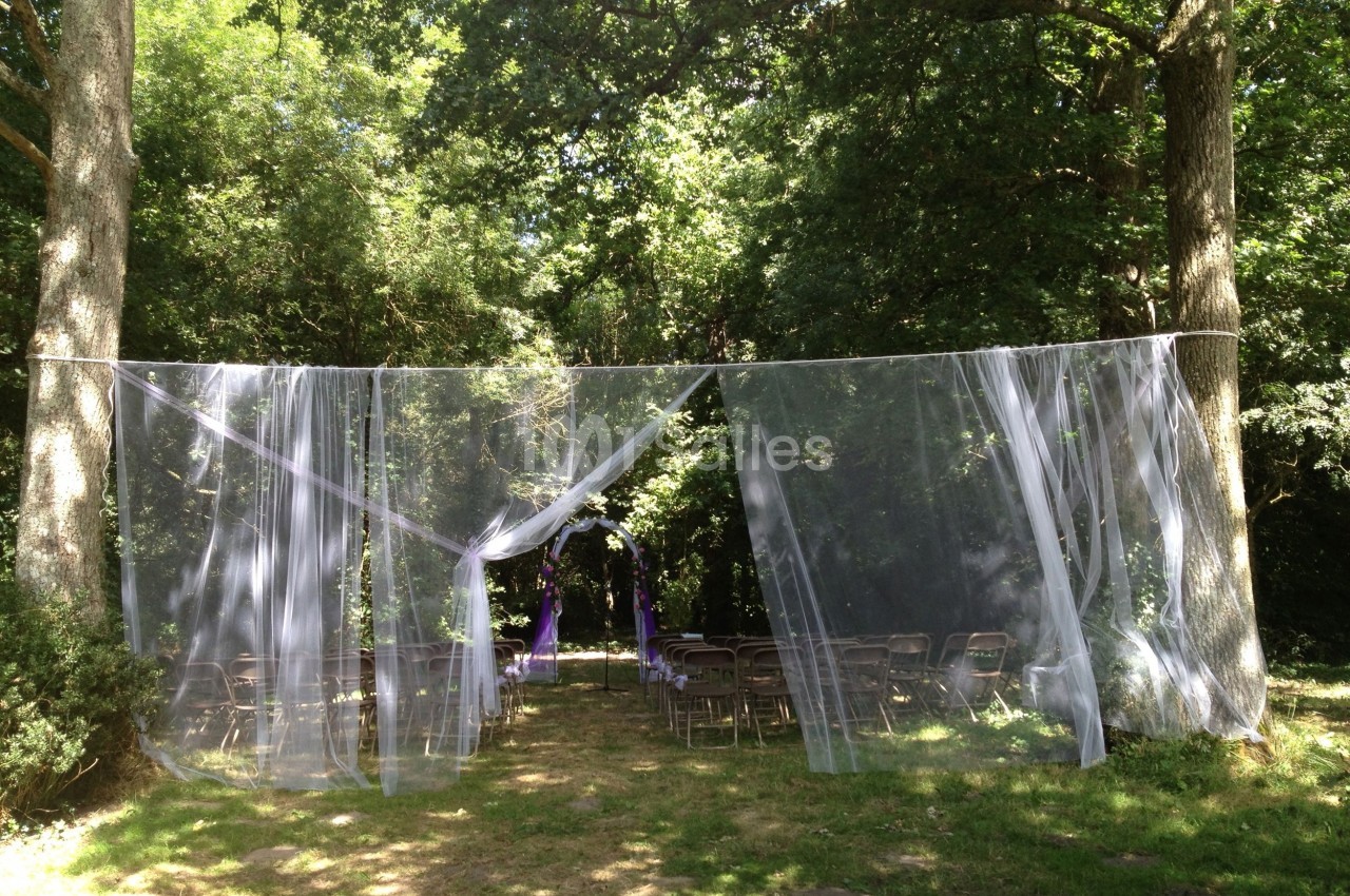 Chaises disposées en extérieur sous des arbres, avec un voile blanc suspendu formant une arche pour une cérémonie.