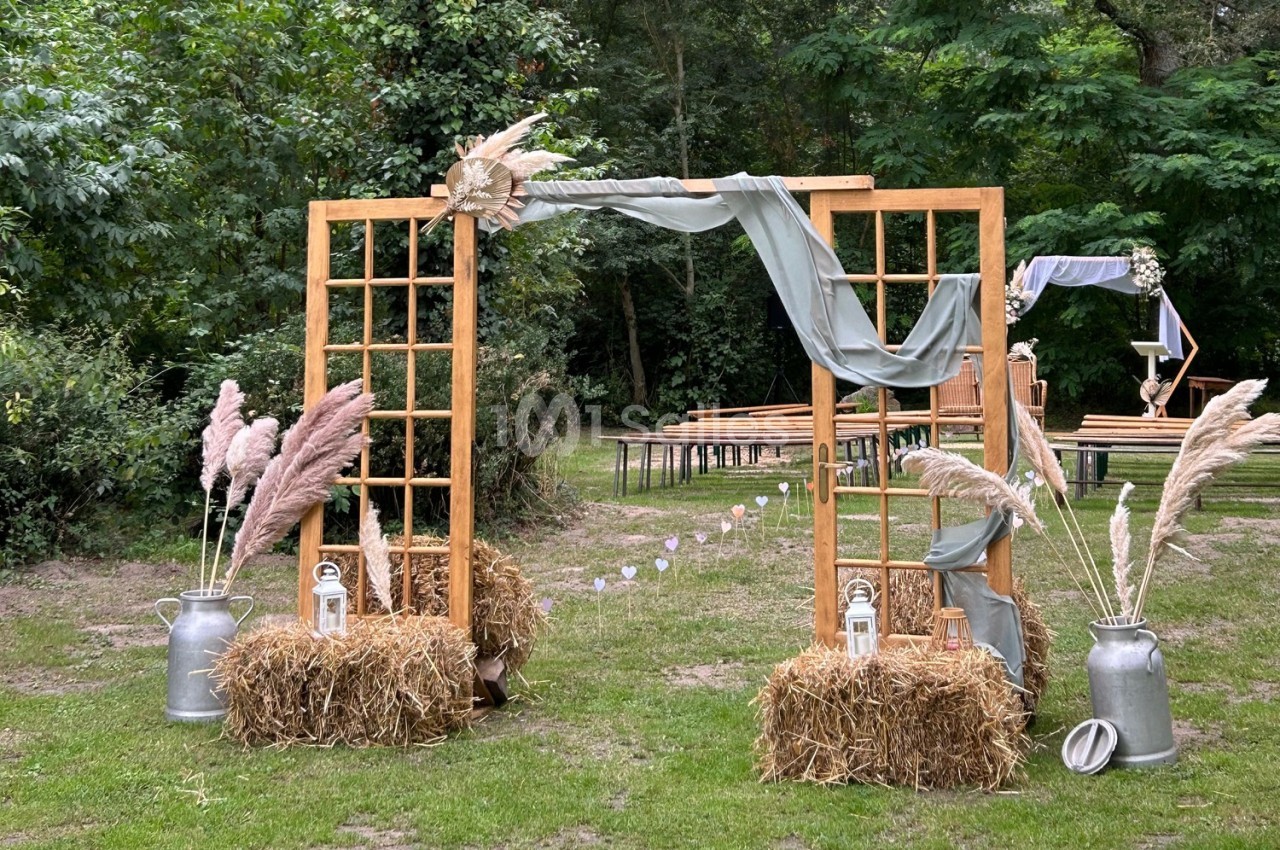 Arche en bois décorée de voilages, pampas et lanternes, installée dans un jardin pour une cérémonie en plein air.