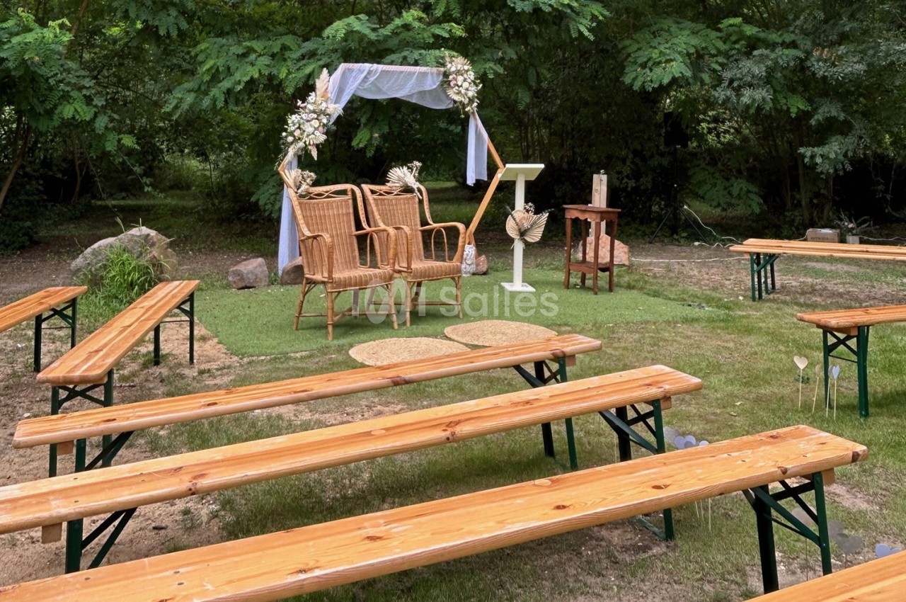 Chaises en osier et arche décorée pour une cérémonie en plein air, entourées de bancs en bois sur une pelouse.