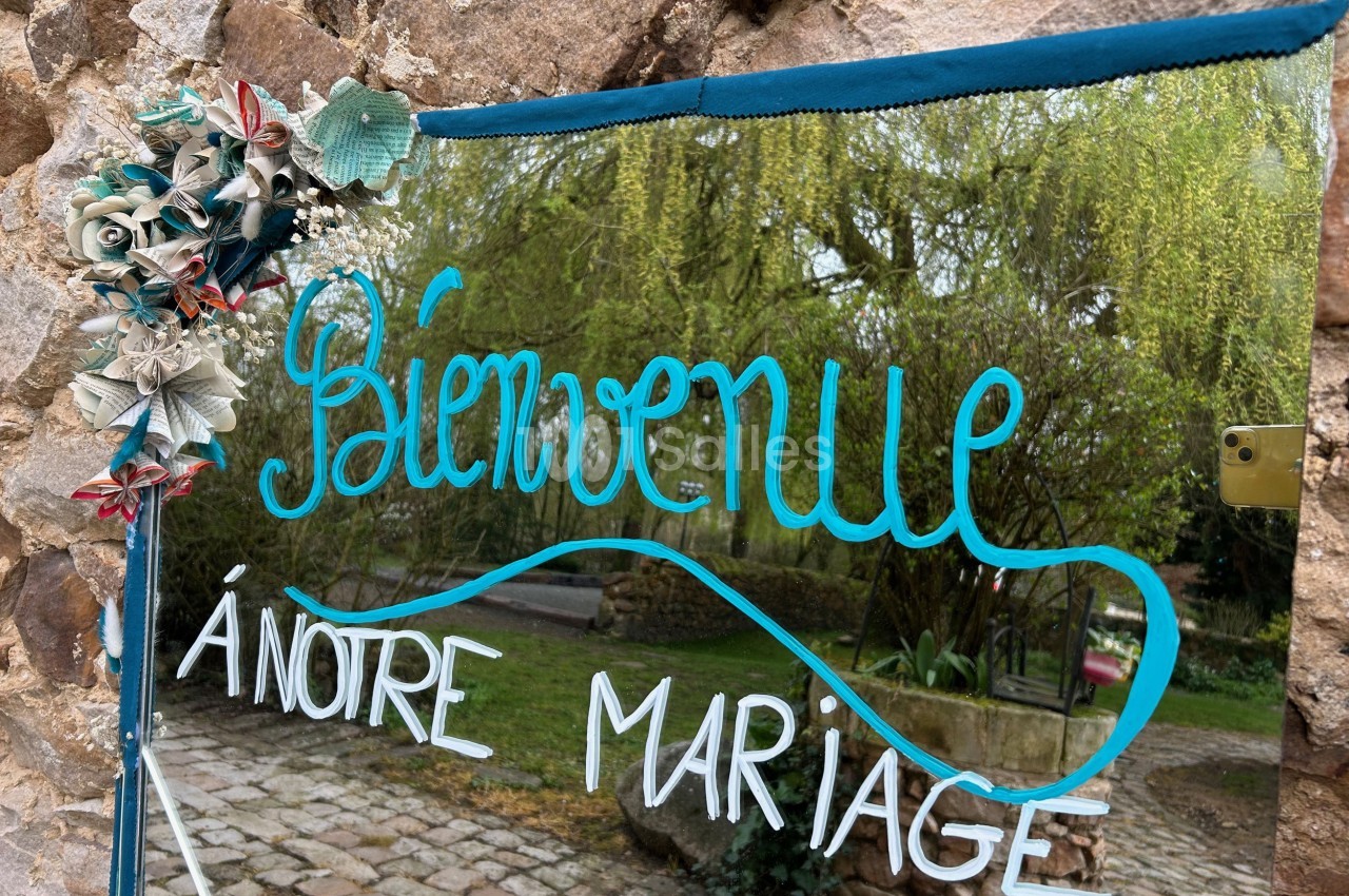 Texte ’Bienvenue à notre mariage’ écrit sur un miroir décoré de fleurs, reflétant un jardin verdoyant.