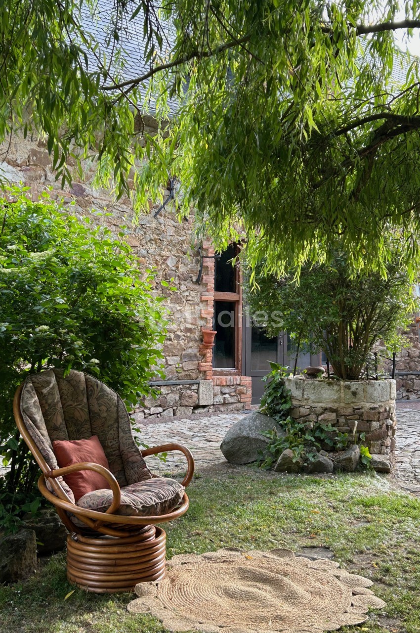 Fauteuil en rotin avec coussins sous un arbre, près d'un mur en pierre et d'une cour pavée.