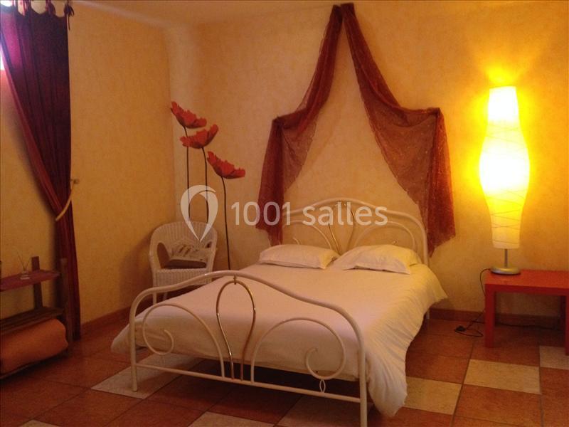 Chambre avec lit double en métal blanc, décoration chaleureuse avec rideaux rouges, lampadaire et fleurs artificielles.