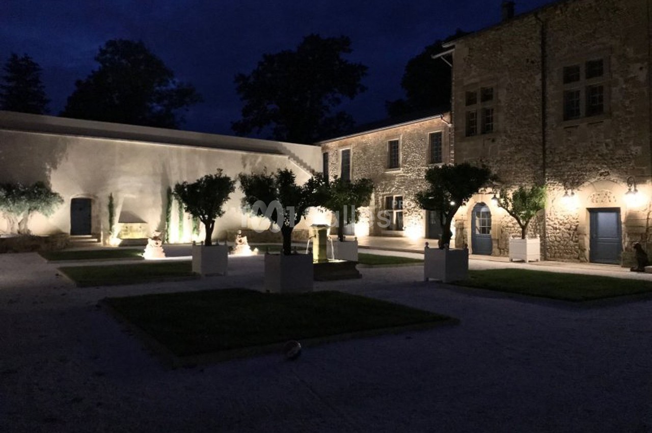 Cour intérieure d'un bâtiment en pierre éclairée la nuit, avec des arbres en pots et un éclairage chaleureux.